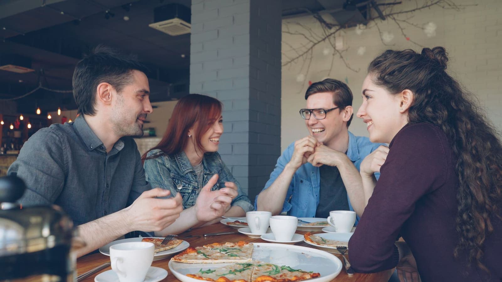 Grupo de amigos a conversar à mesa de um café, com pizza e cafés